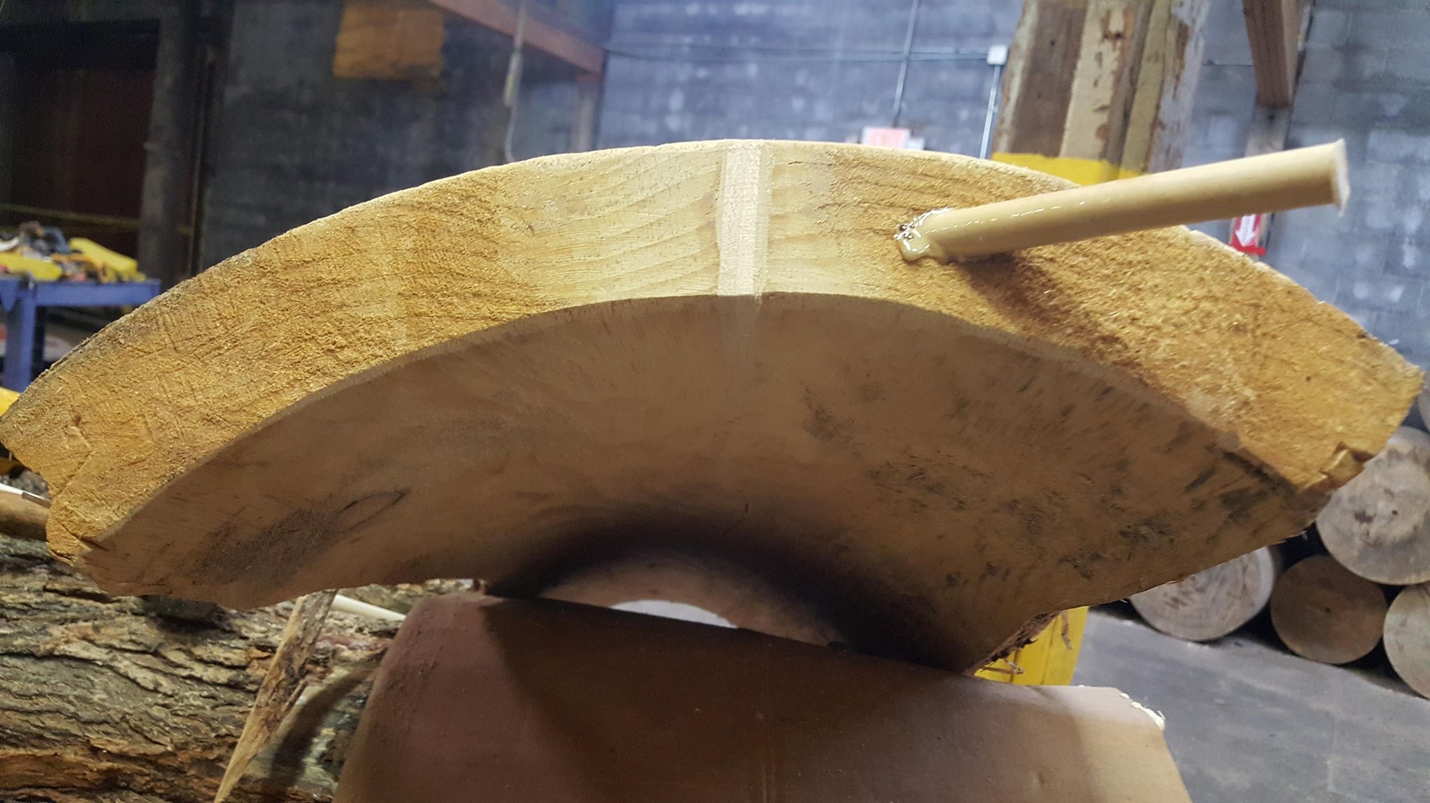 Wide end-on view of the IC WOOD half-log showing the dowel rod spanning the full width of the crack — stacked finished logs visible in background, full geometry of the repair visible